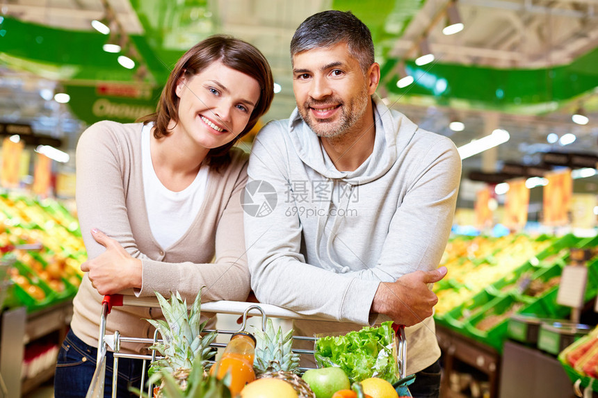 幸福的夫妇在超市里拿着装满产品的购物车看着相机的形象图片
