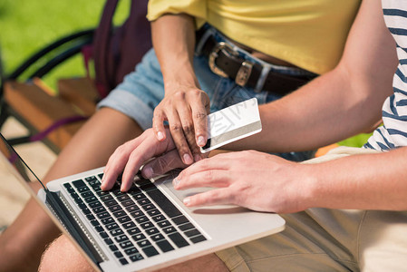 坐在公园的长凳上时用笔记本电脑和信用卡在网上购物的年图片