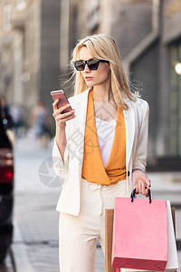 穿着太阳眼镜拿着购物袋和在街上使用智能手机的图片