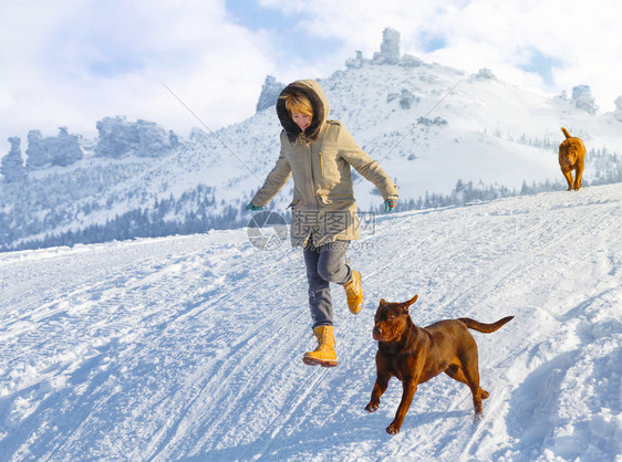 妇女与狗一起在雪田中奔跑和跳跃在阳光明媚的一天对遥远的山峰游戏图片