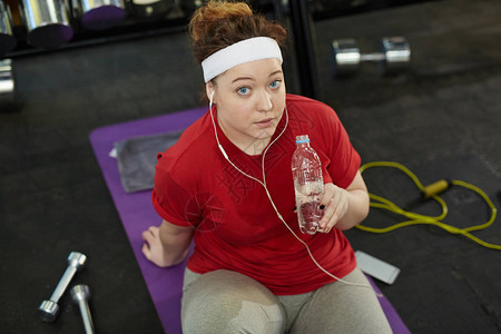 可爱的肥胖女在健身房的垫子上锻炼时做健身运动和听音乐休息喝水图片