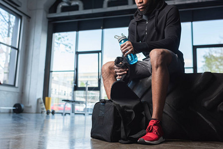 坐在健身房的轮胎上时拿着一瓶水和毛巾的年轻非洲裔美国运动图片