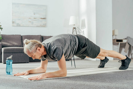 穿着运动服的运动男子在家里的瑜伽垫上做木板运动图片