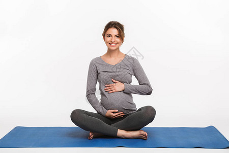 快乐的年轻孕妇坐在瑜伽垫子上抚摸着白图片