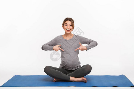 快乐的年轻孕妇坐在瑜伽垫子上指着图片