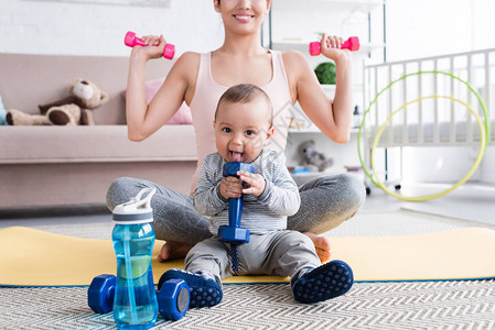 在家和孩子坐在瑜伽垫子上时用哑铃做运动的笑着健康的母图片