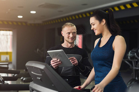 健身教练在椭圆机上帮助年轻女子教练在跑步机上与女孩讨论锻炼计划健身房的有氧运动健康的生活方背景图片