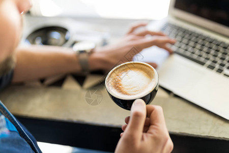 男人喝卡布奇诺咖啡杯和在咖啡店用笔记本电背景图片