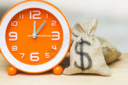 用金钱投资来节省时间和资源的概念美元在钱袋与木桌背景上的白色橙时钟显示货币在图片