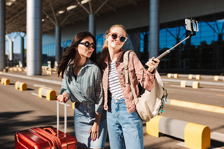 两个戴着太阳镜的漂亮女孩在机场附近的户外肩上带着手提箱和背包在手机图片
