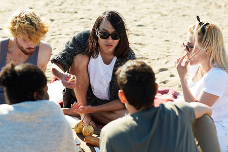 出去玩一群年轻的朋友在沙滩上吃零食和谈野餐背景