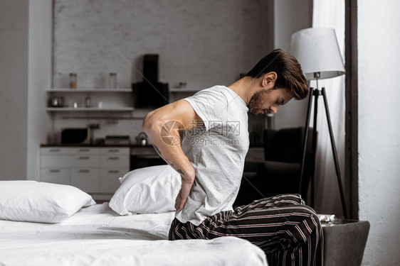 穿着睡衣的青年男子在早晨坐在床上时背痛的侧面图片