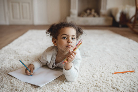可爱的非洲小女孩用铅笔画躺在家里地毯图片