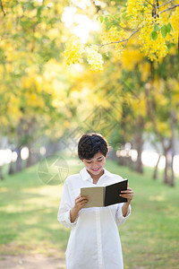 年轻美丽的亚洲女人的肖像站在黄色花朵盛开的公园中夏天季和阅读一本充满图片