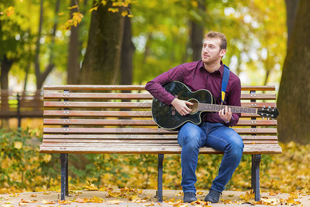 坐在长椅上的年轻人在城市秋季公园弹吉图片