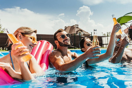 多文化朋友在游泳池中喝啤酒瓶装充气床垫图片