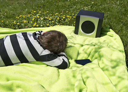 小男孩躺在草地的毯子上听音乐从他的智能手机流到无线和便携式扬声器图片