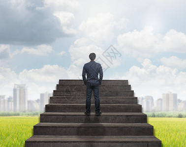 商人站在云天背景下的草地和城市景观上爬楼梯图片