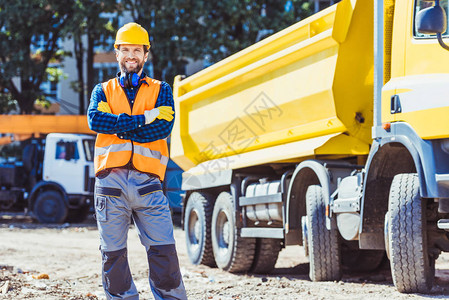 穿着反射背心和硬帽的微笑工人在建筑工地黄色小卡车前用图片