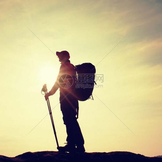 穿着风衣棒球帽和登山杖的徒步旅行者站在山峰岩石上春天阳图片