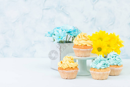 蓝色背景的复古破旧别致花瓶中带有黄色和蓝色奶油装饰和黄色和蓝色菊花束的纸杯蛋糕背景图片