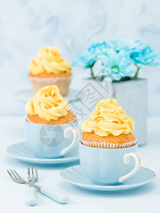 带有黄色奶油装饰的纸杯蛋糕和蓝色柔和背景上复古破旧别致花瓶中的蓝色束图片