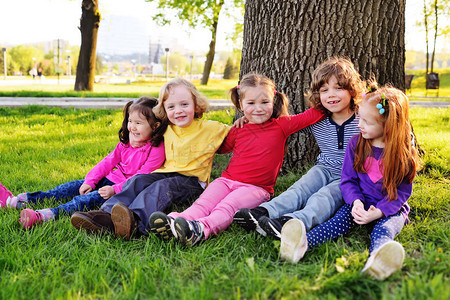 一群穿着五颜六色衣服的小孩拥抱坐在公园里一棵树下的草地上笑着图片