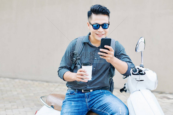 笑着的年轻人坐在摩托车上喝着咖啡杯和在智能手机中检查通知图片