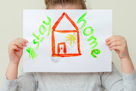 孩子的手拿着一张红房子的照片的特写镜头和待在家里的字样遮住她的脸待图片