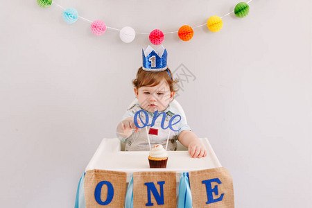 生日快乐派对在家庆祝第一个生日的蓝色冠的逗人喜爱的白种人男婴在高脚椅上蹒跚学步的孩子吃着带字礼帽的美味蛋糕甜图片