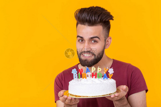 积极英俊的年轻时髦男子在勃艮第t恤拿着祝贺蛋糕与题字生日快乐摆在黄色背景上祝贺和周图片