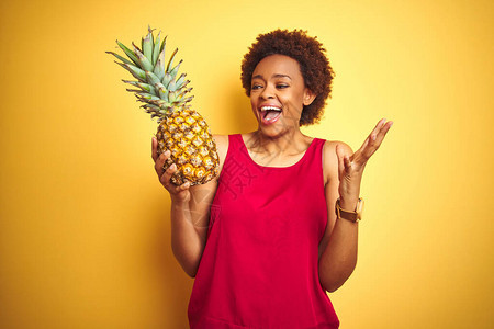 非洲裔美国妇女拿着热带菠萝在黄色孤立的背景下非常高兴和奋图片
