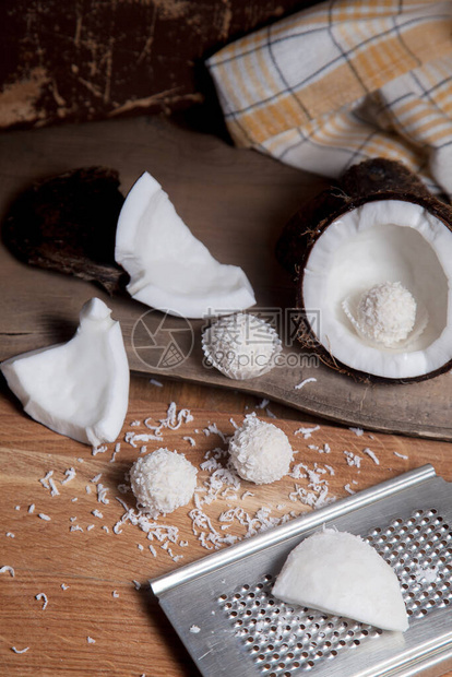 关闭与白色果肉椰子片和甜巧克力椰子松露的椰子椰子壳椰子片碎坚果小块带有椰子片的刨丝器和木制背景上的白巧图片