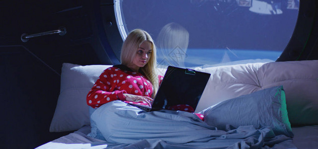 一名年轻女坐在太空船上的床上时使用笔记本电图片