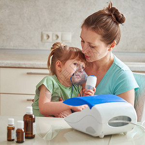女人在家里给孩子吸入将雾化器面罩带到他的脸上吸入药物的蒸气女孩通过面具呼吸桌子上的药背景图片