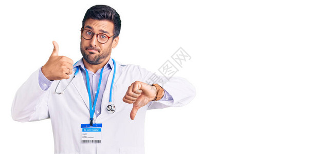 身着医生制服和听诊器的年轻西班牙男子上下伸拇指持不同意见和协议表达意见图片
