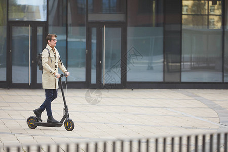 现代年轻商人骑电动滑板车在背景中与城市建筑通勤时的广角肖图片
