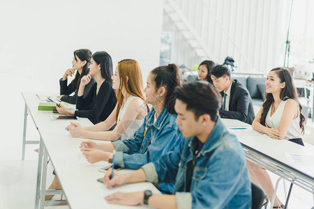 亚洲人参加研讨会并在培训室聆听演讲者的演讲有些人会记笔有人举手询问解说员并在演讲背景图片