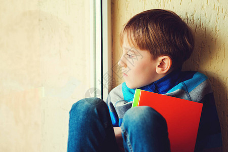 悲伤的男孩坐在窗罩上孩子望着窗外有书的哀伤的男小学生靠近窗户的体贴的孩子独自伤心的孩子家暴负面情绪抑郁图片