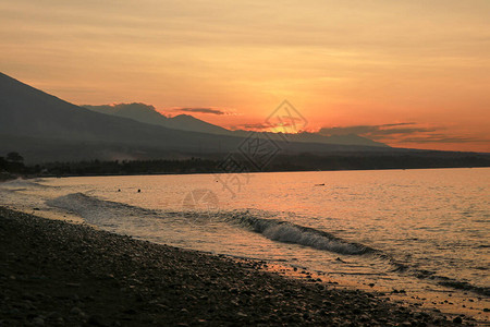 热带岛屿的橙色日落太阳落日照亮的平静海火山和脉全景渔船在海滩上停靠图片