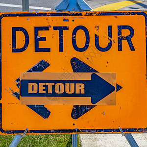 方形关闭一个被风化的橙色和蓝色绕道标志与箭头在路标后面可以看到阳光照射的铺砌道背景图片