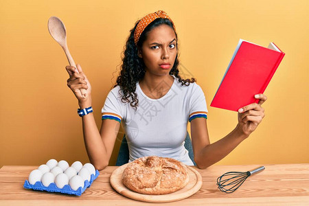 年轻非裔美国女孩做自制面包拿着勺子和书食谱怀疑和紧张图片