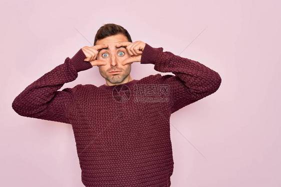 蓝眼睛的年轻帅哥穿着休闲毛衣站在粉红色的背景上试图用手指睁开眼睛图片