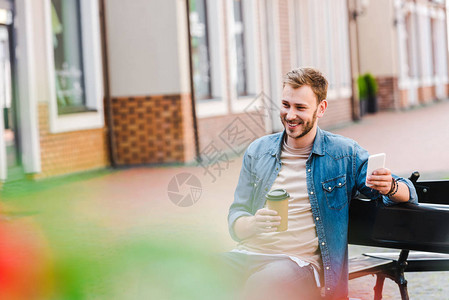 坐在长凳上时拿着纸杯和智能手机的快乐男子有图片