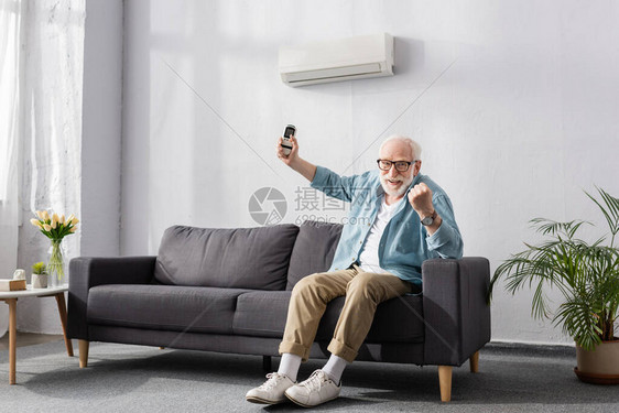 微笑的老人在照相机和沙发上将空调遥控器制器举起手势时表示图片