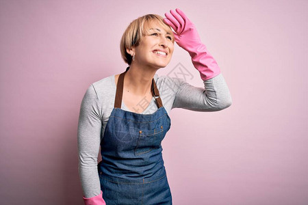 短发穿围裙和手套的金发年轻清洁女工在粉红色背景上非常快乐图片
