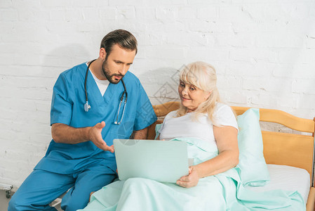 在医院床上使用膝上型看老年妇女的男护士有听诊背景图片