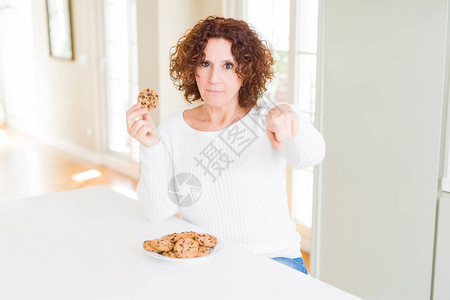 在家里吃巧克力薯饼的老女人用手指着相机和你手牌正面的积极和自信的姿态图片