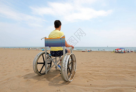 残疾男孩坐在特质轮椅面向大海的沙滩上图片
