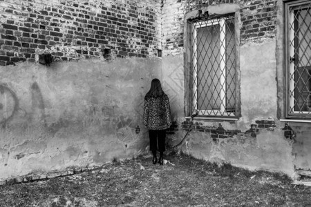 一个女孩背着她站在一栋旧建筑墙边和有铁条的Windows墙上隔离和怨恨图片